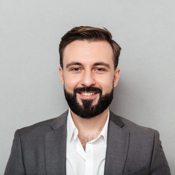 Close up portrait young bearded man white shirt jacket posing camera with broad smile isolated gray 600x600